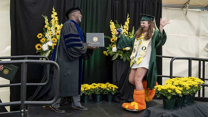 Student wearing the Duck feet walking across the UO Grad stage