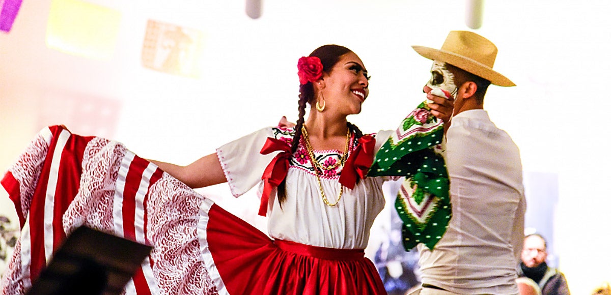 Two dancers in traditional latino dress for a Day of the Dead celebration. Woman in a long red skirt and a man in a man in a white shirt and cowboy hat.