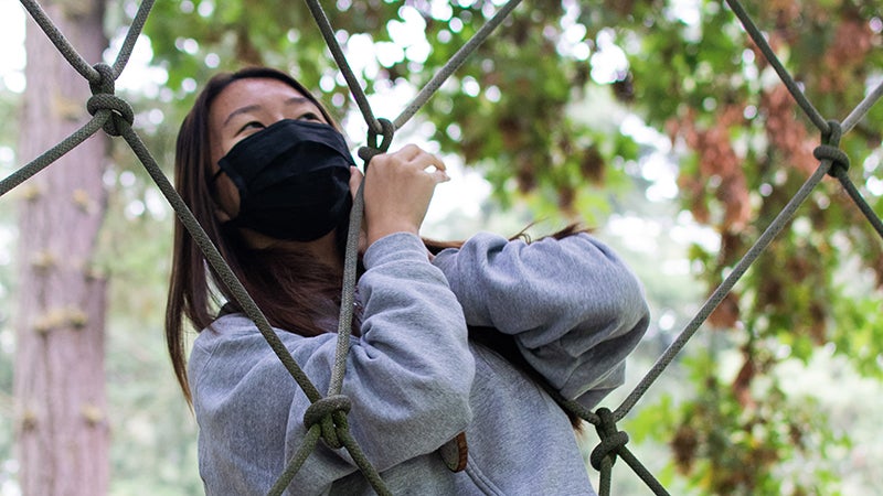 student wearing a mask and holding onto a part of a low ropes course obstacle