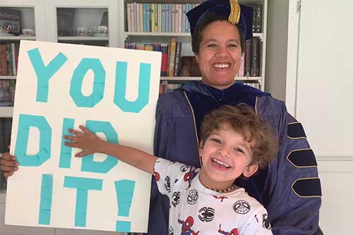 A professor wearing commencement regalia with their child holding a sign that says You Did It!