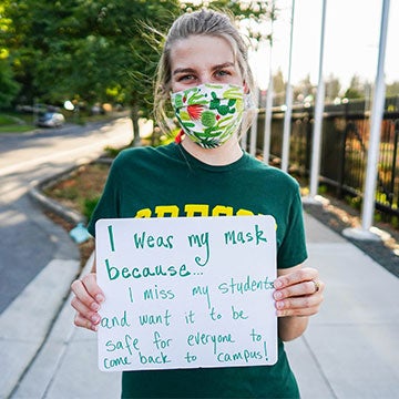 Emily Henkelman wearing a mask with a sign that says I wear a mask because I miss my students and want it to be saf for veryone to come back to campus!