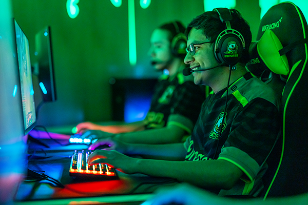 two UO esports players sit at computers in a room lit vibrant green