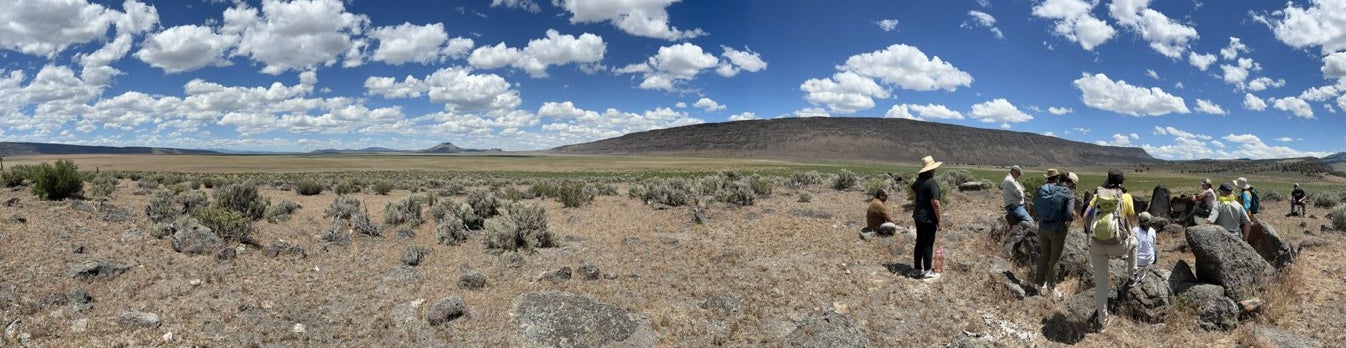 Panoramic view of the Great Basin with a group of people on the right side. There is a bluff in the distance and sage brush on the ground. 