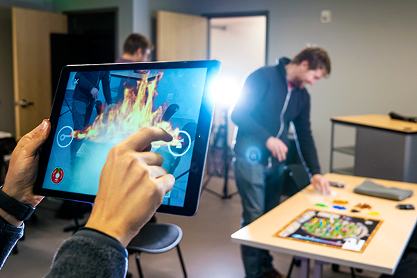 a person clicks on immersive elements on an ipad while another person in the background sets up an immersive game board