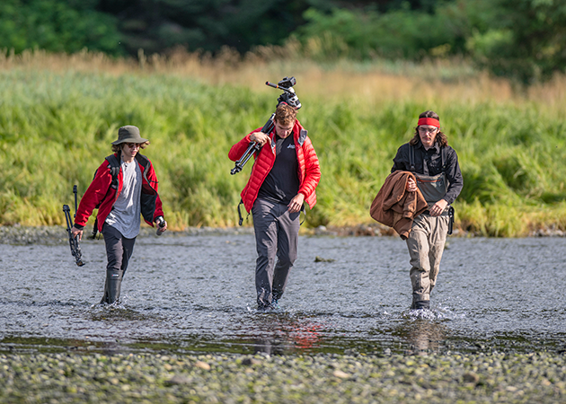 three SOJC students carrying recording equipment walk through a shallow stream