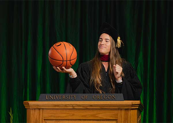 Sabrina Ionescu wearing her commencement regalia and holding a basketball behind a University of Oregon podium