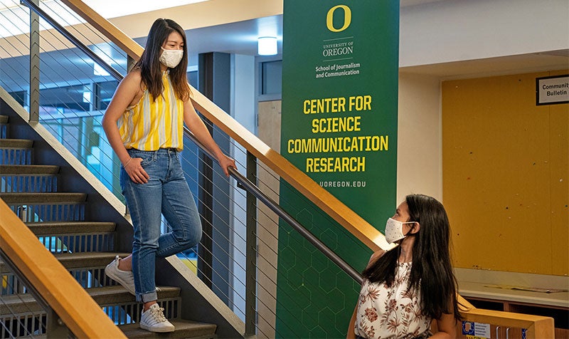 A student wearing a mask walking down the stairs toward another student wearing a mask in Allen Hall with the SOJC Center for Science Communication Research banner behind them