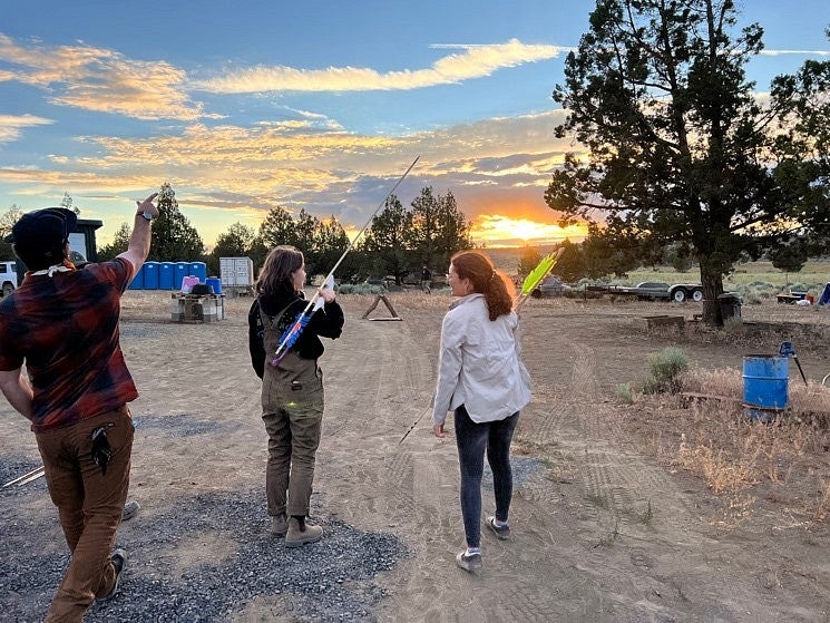 A group of people throw atlatls at camp at sunset.
