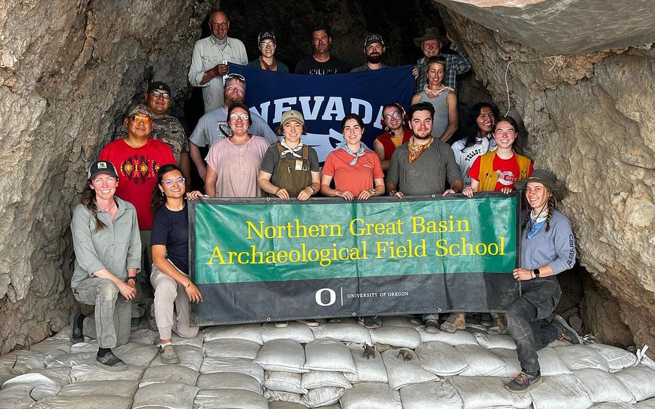 A group of people stand at the back of a cave with a banner reading Northern Great Basin Archaeological Field School.