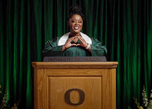 Sabinna Pierre wearing her commencement regalia throwing the O behind a University of Oregon podium