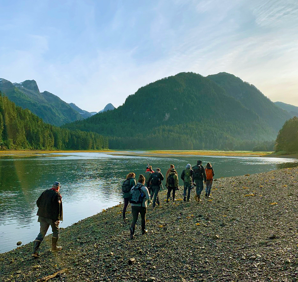 a group of students walk away from the camera along a river bank with forested hills in the background