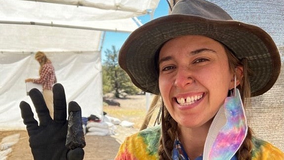 Sonya Sobel, a young woman with a big smile, wearing a tie dye shirt, a tie dye mask hanging from one ear, and a gray hat, holds up an obsidian point.